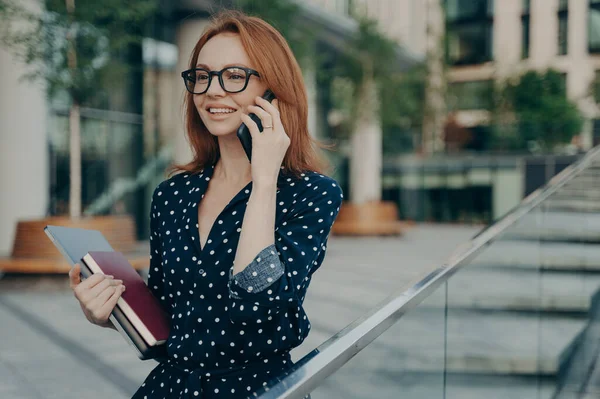 積極的な若い赤毛の女性は 遠くに見える近くにスマートフォンを保つ携帯電話を楽しんでノートパソコンを保持し 日記を身に着けています眼鏡ポルカドットドレスストリートで会うのを待っています — ストック写真