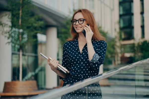 赤い髪をした幸せな大人の女性は 国際話をするためのスマートフォンの支出のレジャーを呼び出すことができます眼鏡ポルカドットドレスは 前方に焦点を当てた都市の設定で屋外で日記ポーズを保持 — ストック写真