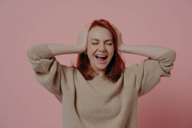Unbelievable! Excited caucasian red-haired woman reacting positively on good news isolated over pink background, overjoyed female with closed eyes holding head in hands and screaming in happiness