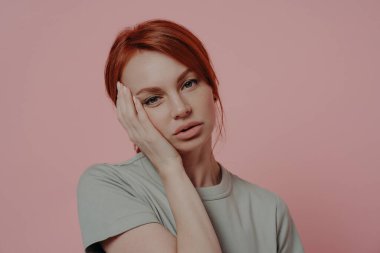Yüzüne elleriyle dokunan, baş ağrısı ya da migrenden muzdarip, pembe arka planda izole edilmiş, sıradan bir tişört giymiş, yorgun ve bitkin bir kızıl kadının portresini yakından çek.