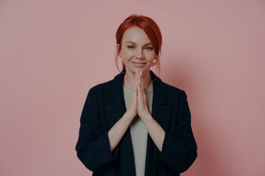 Wishful young red-haired stylish woman dressed in navy casual jacket holding hands in prayer gesture, making wish for good luck, ginger female asking help while posing isolated on pink background