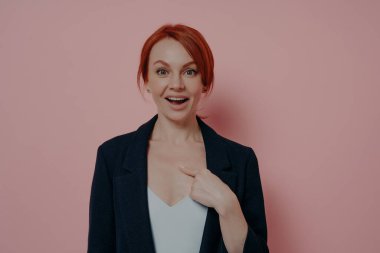 Portrait of happy smiling young red-haired woman asking question you mean me with surprised amazed face expression, keeping mouth opened and pointing at herself, isolated on pink wall background