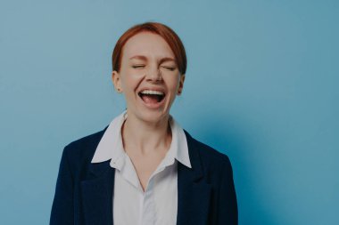 Studio shot of laughing happy woman in formal outfit with closed eyes standing isolated on blue wall, ginger female having fun, hearing funny anecdote or joke from friend. Positive emotions concept