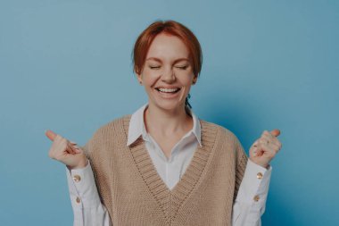 I did it. Studio portrait of overjoyed happy excited red haired female raising clenched fists and keeping eyes closed, isolated on blue background, hearing amazing news. Success concept