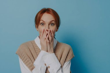 Surprise. Young shocked amazed redhead woman in stylish outfit covering mouth with both hands and being surprised, hearing unexpectable news from friend, isolated over blue background