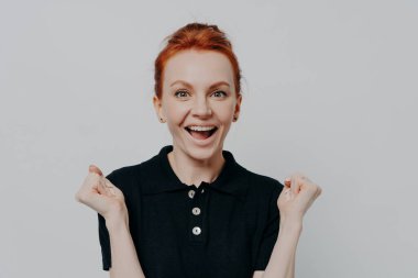 Siyah polo tişörtlü mutlu genç kız harika haberlerle heyecanlanıyor. Ağzını açık tutuyor ve yumruklarını sıkıp ellerini kaldırıyor. Zaferi kutluyor.