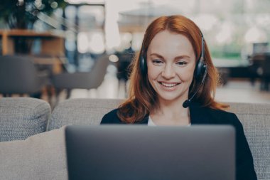 Kızıl saçlı güzel girişimci kızın yakın plan portresi, uzaktan bilgisayar ve kulaklıkla çalışıyor, müşterilerle internet üzerinden iletişim kuruyor, müşteri hizmetleri raporlarını ve sık sık taleplerini kontrol ediyor.