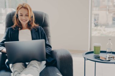Modern dizüstü bilgisayarla çevrimiçi konferans sırasında mutlu gülümseyen kadın serbest yazar, kulaklıklı neşeli kızıl iş kadını evde koltukta dinlenirken webinar ya da eğitim videosu izliyor.