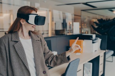 Kızıl saçlı, VR kulaklık takan, parmaklarıyla bir şeye dokunan, sanal gerçekliği ilk kez deneyen, modern ofis ortamında mobilyaları bulanık arka planda duran genç bir kadın.