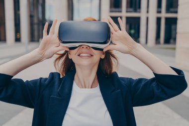 Genç, gülümseyen kızıl saçlı kadın girişimci taşınabilir VR gözlükleri test ediyor, iş kadını sanal gerçeklikle etkileşime geçiyor ve şehirde tek başına dururken olumlu duygular ifade ediyor.