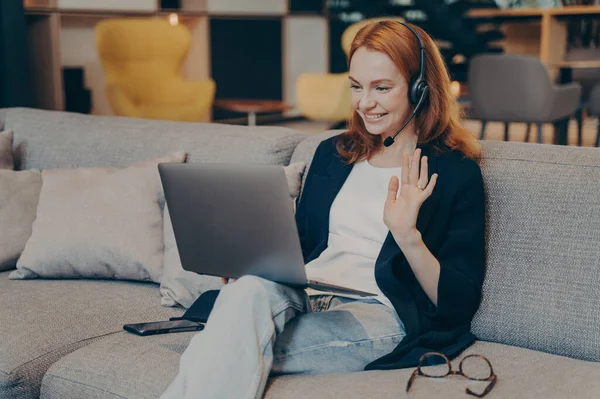 友人とインターネットビデオ通話のためのラップトップと無線ヘッドセットを使用して若い女性は 挨拶ジェスチャーで手を振って 広くコンピュータ画面を見ながら笑顔 オンライン通信の概念 — ストック写真