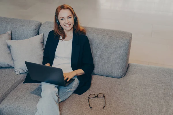 Beautiful young woman with red hair in casual clothes sitting with laptop and wireless headset on cozy grey sofa in public place, looking up at camera with beaming smile, working distantly online
