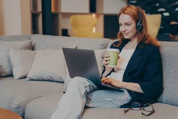 若い赤毛の女性は ノートパソコンやワイヤレスヘッドセットのイヤフォンを介してオンラインで友人とチャットしながら 現代のコーヒーハウスのインテリアでソファに座って 彼女の手でドリンクとカップを保持 — ストック写真