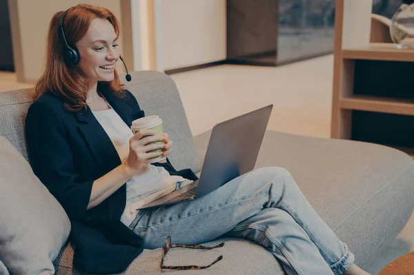 ノートパソコンでオンラインのウェビナーコースを見ながらヘッドフォンと笑顔を身に着けている幸せな千年紀の女性は 地元の茶店で居心地の良いソファに座ってコーヒーを飲みました 遠隔Eラーニングの概念 — ストック写真