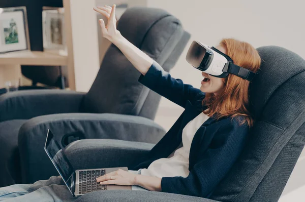 Excited woman in VR headset and laptop, touching air during virtual reality experience while sitting in chair in internet cafe salon. Emotional lady exploring artificial 3D world or playing video game