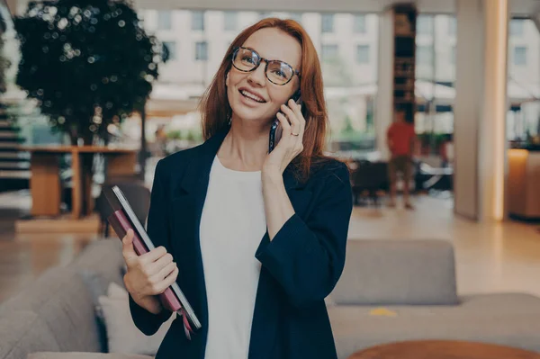 ノートパソコンとノートパソコンを手にした成功したジンジャー女性起業家を自信を持って クライアントと携帯電話で楽しい会話を楽しみ 眼鏡と正式な服を着て — ストック写真