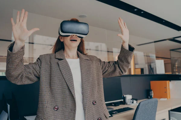 Woman with surprised facial expression experiencing amazing world of virtual reality with VR goggles, standing alone in front of office desk, holding her hands in air, using 3d glasses at work