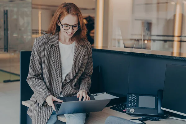 Young beautiful female office employee in casual clothes at work, sitting on top of desk, using laptop to browse files, redhead smiling businesswoman preparing for presentation while working in office