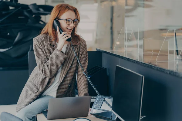 Gözlüklü bir kadın ofiste çalışıyor, günlük kıyafetler giyiyor, masasında otururken telefonla konuşuyor, modern ve rahat ofis ortamı, bulanık arka plan. İş konseptinde mutlu çalışan