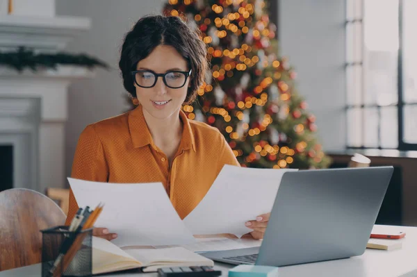 Smiling Hispanic woman employee working in office at christmas time, female checking invoice documents and company reports for year-end financial meeting sits at desk with laptop against New Year tree