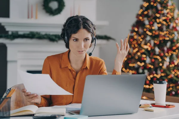 Serious spanish woman teacher in wireless headset with microphone giving online lesson on laptop while working at home office on Christmas, sitting in room with decorated xmas tree on background