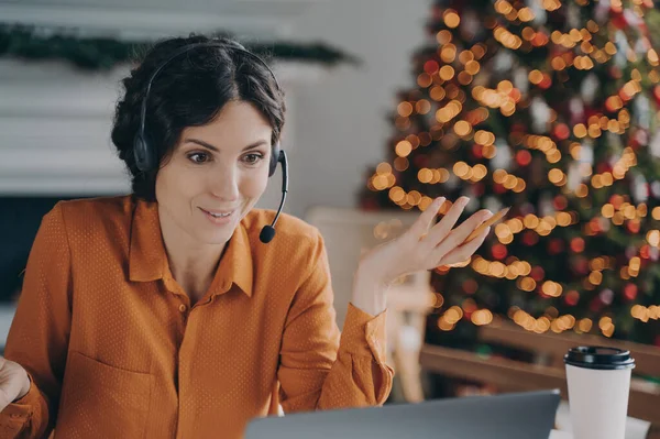 Helpful call center consultant beautiful hispanic woman in headset with mic communicates with customer while working remotely online during Christmas holidays, sitting at desk against xmas tree