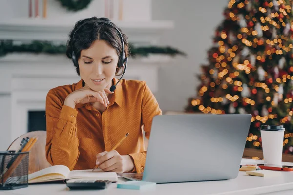 European female call center oparator in headset with mic making notes in agenda while speaking with client via laptop online, works from home with Xmas tree on background. Work during winter holidays