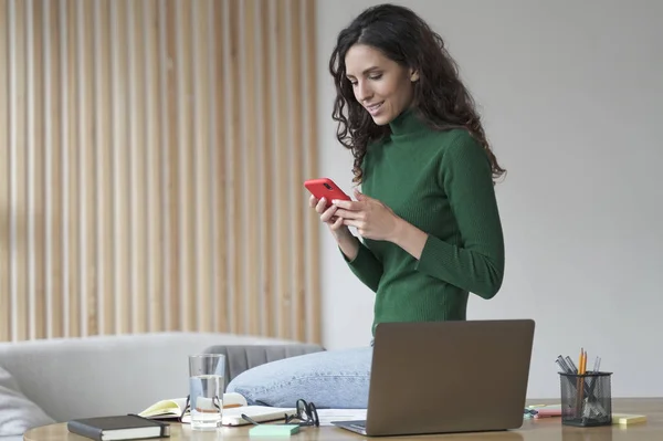 Young happy italian woman freelancer using mobile application at her workplace with laptop computer during remote work at home, spanish female reading online news or sending email from smartphone