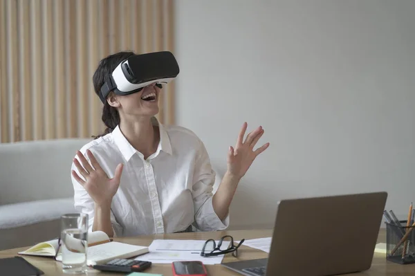 Enthusiastic business lady wears VR headset for laptop raising hands up as trying to touch objects in 3D reality when sitting at desk, working with apps online, modern technology for business concept