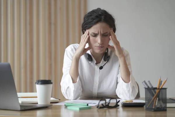 Stressed young entrepreneur lady suffering from headache massaging temples while sitting at home office, tired female freelancer feeling unwell, touching head. Migraine pain, fatigue, stress concept