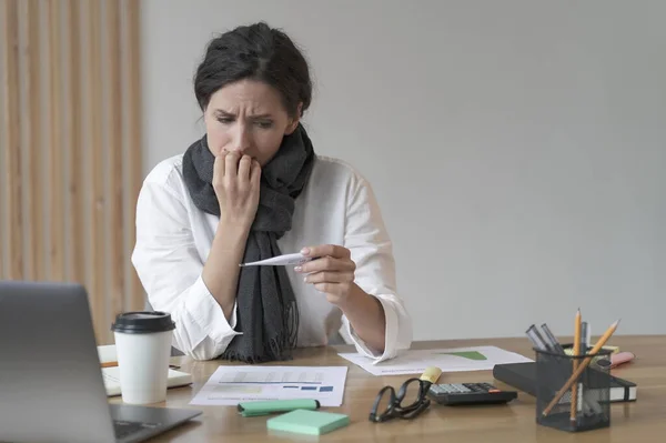 Unhappy latin business woman dressed in warm scarf checking body temperature at work, looking at thermometer with worried face expression, suffering of covid or flu symptoms at workplace