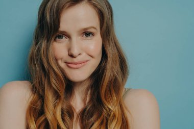 Çekici, utangaç, kahverengi saçlı ve kameraya bakan genç bir kadının çekimleri. Mavi arka planda izole edilmiş çıplak omuzlu ürkek bir kadının portresi. Kadın güzellik konsepti