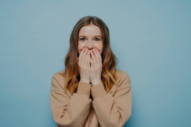 Bej renkli süveter giymiş genç bir bayan, olaya ya da haberlere şaşırıyor. Ağza karşı el ele tutuşuyor ve mavi arka planda izole edilmiş, şok olmuş yüz ifadesiyle kameraya bakıyor.