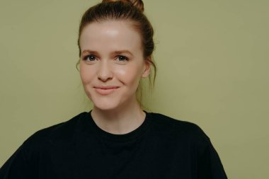 Siyah tişörtlü, siyah tişörtlü genç bir kadının yakın portresi. Hafif bir sırıtışla kameraya bakıyor, gülüşünü tutmaya çalışıyor, açık yeşil arka planda tek başına duruyor.