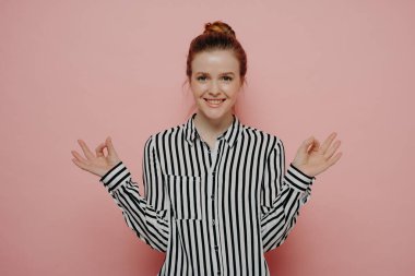 Sakin ve rahat genç kızıl saçlı kadın, günlük kıyafetleriyle iki eliyle ya da meditasyonla iyi bir jest yapıyor, pembe stüdyo arkasında tek başına dururken kendini iyi hissediyor.