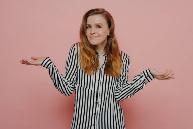 Siyah çizgili beyaz gömlekli genç bir kadın, iki eli havada omuz silkiyor, stüdyoda pembe duvarın yanında otururken ne yapacağını bilmiyor. Genç kız kararsız görünüyor.