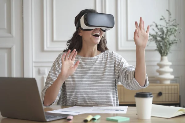 Photo of excited amazed woman wearing VR glasses enjoying amazing virtual reality experience while sitting at office table, making purchases in 3d store. Innovation and technology concept