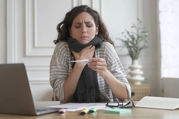 Worried young hispanic woman office worker sitting at table with laptop, looking at thermometer, sick female employee with high fever suffering from cold flu at work, checking temperature at workplace