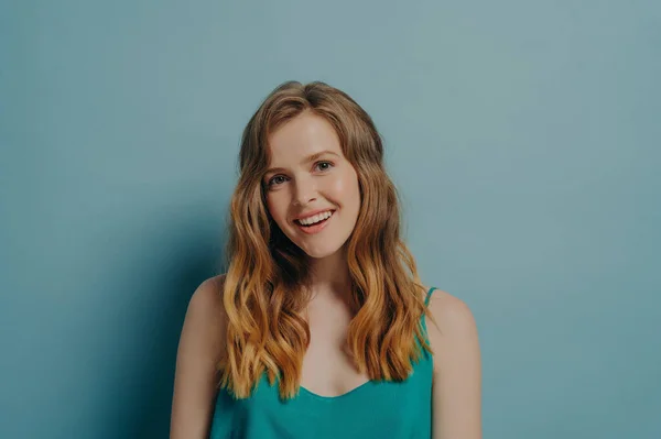 Happy young light brown-haired woman posing with head tilted, looking at camera with gentle smile opening her mouth slightly shows perfectly straight teeth isolated over blue studio background