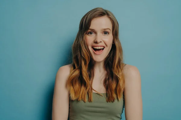 Wow emotion. Studio portrait of amazed astonished young female in tank top expressing happiness and excitement, looking at camera with surprised face expression, standing isolated on blue background