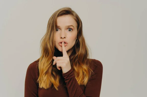 Mysterious caucasian teen girl holding finger on lips and looking at camera with scared face expression, female asking to be silent and making shh gesture while posing isolated on grey background