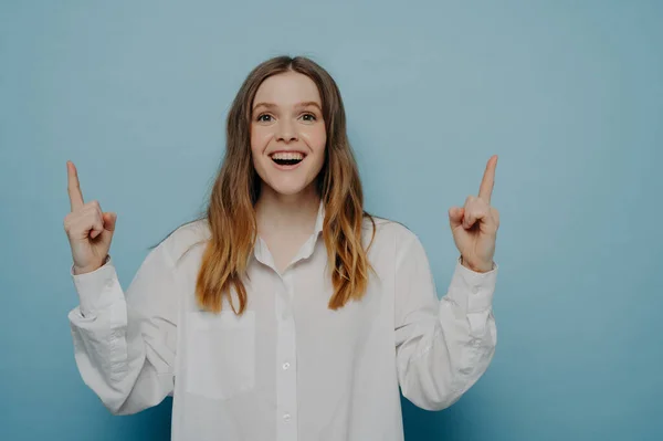 Amazed young woman pointing upwards with fingers and expressing excitement while seeing something amazing, isolated on blue wall, offering great opportunity. Advertisement and promotion concept