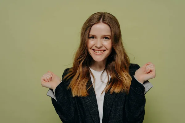 Happy young female with wavy brown hair holding hands up demonstrating excitement wearing dark formal jacket and white top, posing against light studio background. Human emotions concept