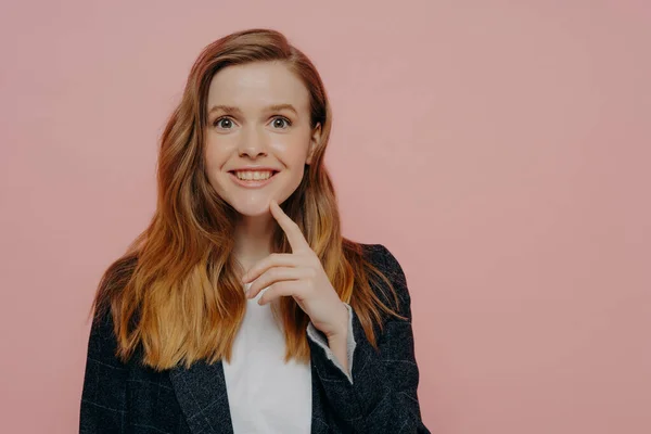 Young excited ginger lady in jacket with white shirt, realizing there is solution, generating new idea with finger on chin, standing alone next to pink background and expressing happiness