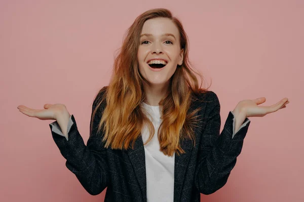 Laughing young woman raising hands using open palm gesture and looking ahead with amusement and surprise wearing dark formal jacket and white top posing against pink wall