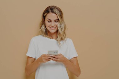 Genç mutlu kız akıllı telefon kullanıyor, sosyal medyada erkek arkadaşıyla sohbet ediyor ve turuncu duvara karşı beyaz tişört giyerek gülümsüyor. Modern teknolojiler ve ağ kurma kavramı