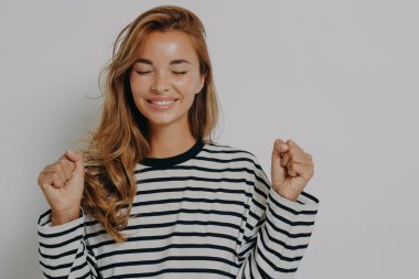 Rüyalar gerçek olacak. Heyecanlı genç bir kadın, şans için yumruklarını sıkıyor, gözleri kapalı gülümsüyor, servet ve başarı bekliyor, şans dileniyor, gri stüdyo geçmişinde izole edilmiş.