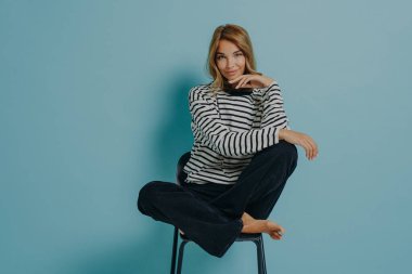 Çekici genç bayan model sandalyenin üstünde oturuyor, bacakları havada ve bir eli çenesinde, rahatlamış hissediyor, ve kameraya gülümsüyor, mavi stüdyo arka planında izole edilmiş.