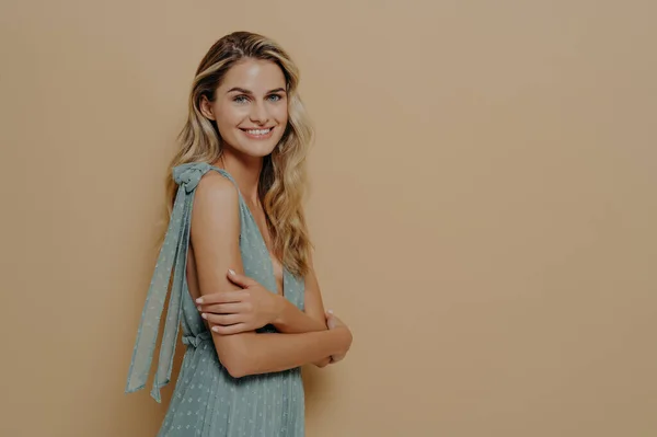 Romantic young and attractive female model in beauriful evening dress getting ready for perfomance standing sideways delighted and happy with crossed hands, smiling joyfully at camera