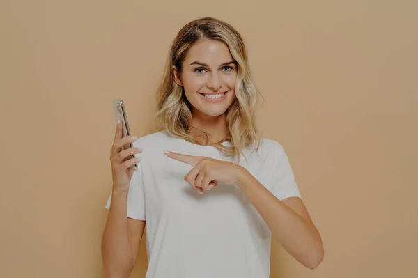 Cute excited female pointing on smartphone with forefinger and looking at camera with broad smile, reading good news in internet and feeling happy while posing isolated over beige background